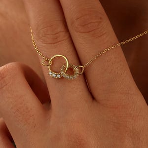 May include: A gold necklace with two linked circles, one set with small, sparkling gemstones. The necklace is shown against a skin tone background, emphasizing the jewellery's intricate details and elegant design.
