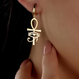 May include: Gold-colored hoop earrings with an Ankh and Eye of Horus pendant. The earrings are made of a shiny, reflective metal and are worn on the ear. The Ankh symbol is a cross with a loop at the top, and the Eye of Horus is a stylized eye.