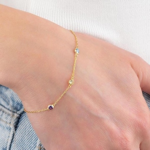 May include: A delicate gold bracelet featuring four colorful gemstones: purple, yellow, blue, and clear. The bracelet is worn on a wrist, with the chain and gemstones visible against the skin. The bracelet is a simple, elegant piece of jewelry.
