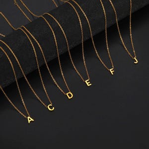 May include: Gold-coloured initial necklaces. Each necklace has a delicate chain and a single letter pendant. The letters A, C, D, E, F, and J are visible. The necklaces are displayed against a dark background.