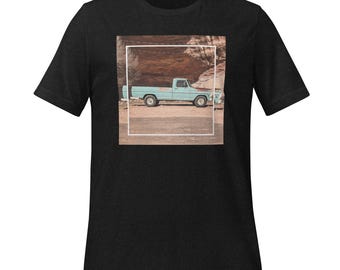 Könnte beinhalten: Schwarzes T-Shirt mit einem quadratischen Bild eines hellblauen Oldtimer-Pickups. Der Truck steht auf einer unbefestigten Straße vor rötlich-braunen Felsformationen. Das Shirt besteht aus einem weichen, dunklen Stoff.