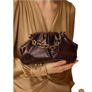 Peut inclure: Une pochette marron métallique foncé avec une texture façon peau de serpent et une silhouette froncée en forme de bourse. Le sac présente une anse épaisse en acrylique à motif écaille de tortue fixée sur le dessus du cadre.