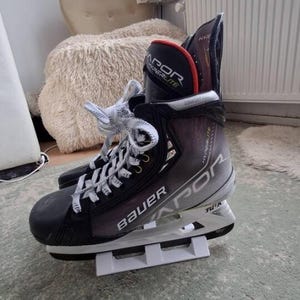 May include: A Bauer Vapor Hyperlite ice skate featuring a black and grey gradient boot with white laces, a black toe cap, and a white Tuuk blade holder. The tongue displays the text Vapor Hyperlite in white and red, with Comfort Edge branding.