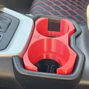 May include: A bright red dual cup holder insert designed for a vehicle center console. The plastic accessory features two circular openings with a cutout design and the text VL030 embossed on the surface, fitting securely into the existing black console.