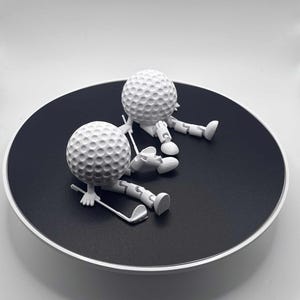 May include: Two white golf ball figures with articulated limbs and golf clubs rest on a black circular surface with a white border. The golf balls have dimpled surfaces, and the figures appear to be in a relaxed pose.