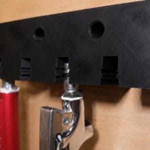 Könnte beinhalten: Eine schwarze Wandhalterung aus Kunststoff mit fünf Schlitzen zur Aufbewahrung von Werkzeugen. Drei Werkzeuge stecken derzeit in der Halterung: ein rotes Werkzeug links, ein metallisch silbernes Werkzeug in der Mitte und ein blaues Werkzeug rechts.