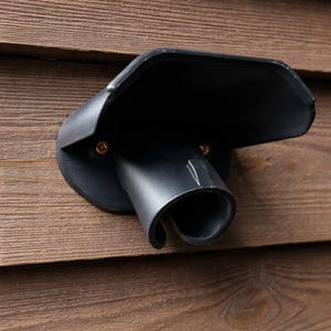 Puede incluir: Una estación de carga negra, montada en la pared, para un vehículo eléctrico. La estación tiene una salida cilíndrica curva y una cubierta protectora. Está montada sobre una superficie de madera con vetas visibles.