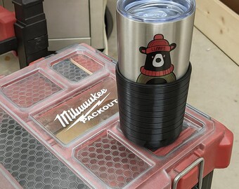 May include: A stainless steel tumbler with a black silicone sleeve, featuring a cartoon bear wearing a red hat and scarf. The tumbler is resting on a red and black toolbox with the "Milwaukee PACKOUT" logo.