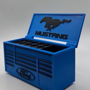 Puede incluir: Una caja de herramientas azul con detalles negros y el logotipo de Ford Mustang. La caja de herramientas tiene una tapa con bisagras y compartimentos internos. El logotipo de Ford está en la parte delantera y el logotipo de Mustang en la tapa.