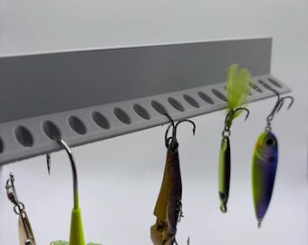 May include: A gray fishing lure holder with several fishing lures attached. The lures are various colors, including gold, green, and purple. The holder has multiple holes for the lures to hang from. The background is plain white.