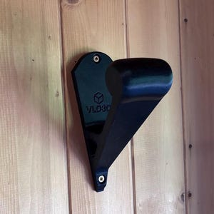 Puede incluir: Un gancho de pared negro, impreso en 3D, montado sobre una superficie de madera. El gancho tiene una parte superior curva y una base triangular, con el texto "VLD3D" visible. Fijado con tornillos.