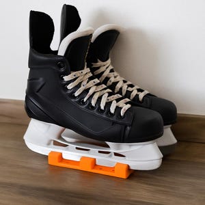 Könnte beinhalten: Ein Paar schwarze Eishockeyschuhe mit weißen Schnürsenkeln und einem weißen und orangefarbenen Klingenschutz. Die Schlittschuhe stehen vor einer weißen Wand und einem Holzboden.