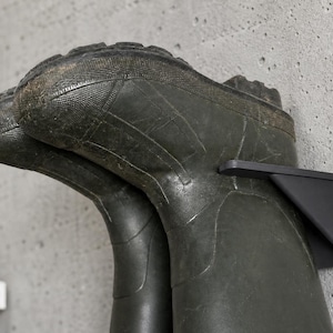 May include: A pair of dark green rubber boots hanging on a black metal wall-mounted boot rack. The boots are textured and show signs of wear. The rack is mounted on a gray concrete wall.