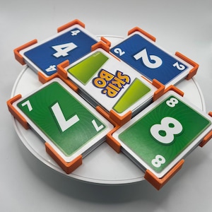 Peut inclure: Un ensemble de cartes du jeu Skip-Bo, avec des cartes bleues, vertes et blanches portant de grands chiffres. Les cartes sont maintenues par des supports orange. Le logo Skip-Bo est visible sur une carte.