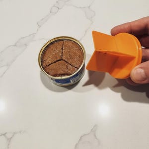 May include: An open can of pet food with a brown, textured interior, divided into three sections. An orange plastic can opener is positioned next to the can, ready for use. The can has a gold rim and a blue and white label.