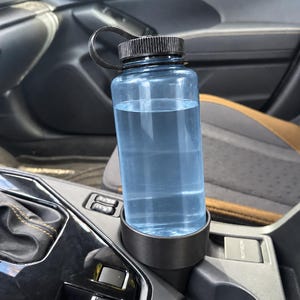 May include: A translucent light blue plastic water bottle with a black screw-top lid and a flexible black loop handle. The bottle is filled with clear water and is securely positioned inside a black cylindrical cup holder insert.