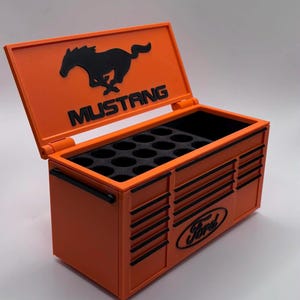 Puede incluir: Una caja de herramientas naranja con detalles negros y el logotipo de Mustang en la tapa. El logotipo de Ford está en la parte delantera. La caja de herramientas tiene un interior negro con múltiples compartimentos.