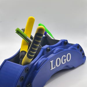 Puede incluir: Un organizador de escritorio azul con forma de pinza de freno de coche, que contiene bolígrafos y rotuladores. La palabra "LOGO" es visible en blanco. Los bolígrafos y rotuladores son amarillos, verdes y negros.