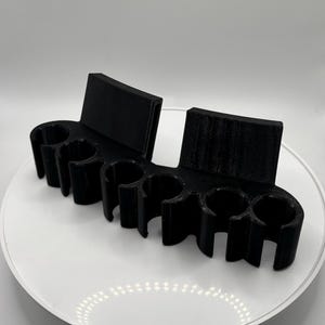 Puede incluir: Un soporte organizador negro impreso en 3D con seis clips circulares dispuestos en fila, diseñados para sujetar objetos cilíndricos. La unidad incluye dos soportes de montaje planos y rectangulares en la parte posterior para estabilidad.