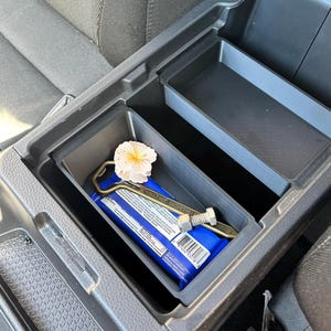 May include: A black plastic vehicle center console storage compartment containing a blue rectangular package, a metallic bottle opener with the text THE NORTH, a small silver metal bolt, and a white artificial flower with a yellow center.
