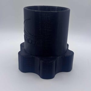 Puede incluir: Un objeto cilíndrico negro con una base en forma de estrella. El objeto tiene una superficie texturizada y un logotipo en el lateral. El diseño parece estar impreso en 3D. El objeto es probablemente un contenedor o soporte.