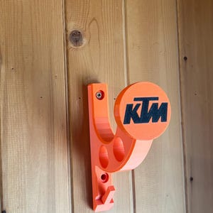 May include: An orange wall-mounted holder featuring the KTM logo in black. The holder is made of plastic and has a curved design with two circular cutouts. It is mounted on a wooden wall.