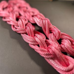 May include: Pink braided rope with a decorative knot pattern. The rope is made of a soft, flexible material and has a subtle sheen.