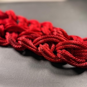 May include: A close-up of a red rope woven into a complex pattern. The rope is soft and pliable, and the intricate design is highlighted by the light reflecting off the surface.