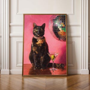 Op de afbeelding: Een ingelijst schilderij van een kat met een juwelen halsband, zittend voor een roze achtergrond. Een martini glas met een olijf staat voor de kat, en een discobal is op de achtergrond. De kat heeft zwart en bruin haar.