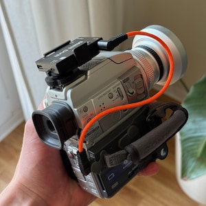Könnte beinhalten: Silberner Sony Camcorder mit einem schwarzen Mini-DV-Adapter und einem orangefarbenen Kabel. Der Camcorder hat einen schwarzen Sucher, einen grauen Griff und verschiedene Knöpfe. Auf dem Mini-DV-Adapter steht "MINI DV".