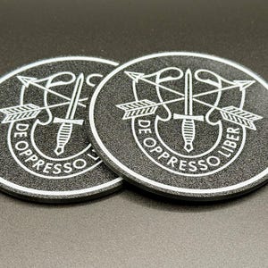 Op de afbeelding: Twee zwarte onderzetters met een witte afbeelding van het insigne van de 101e Airborne Division, met een dolk en pijlen, en de tekst "De Oppresso Liber".