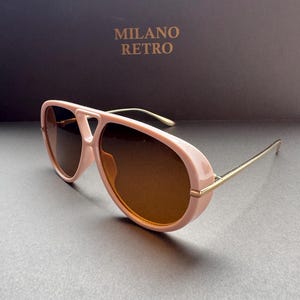 May include: Pink aviator sunglasses with brown lenses and gold-colored temples. The sunglasses have a retro design. The text "MILANO RETRO" is visible in the background.