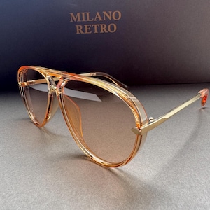 Op de afbeelding: Retro zonnebril met een lichtbruine tint en goudkleurige monturen. De zonnebril heeft een dubbele brug en een dik, helder plastic montuur. De tekst "MILANO RETRO" is zichtbaar op de achtergrond.
