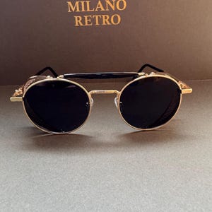 May include: Gold-rimmed sunglasses with round black lenses. The sunglasses have a double bridge and side shields. The text "MILANO RETRO" is in gold lettering on a brown background.