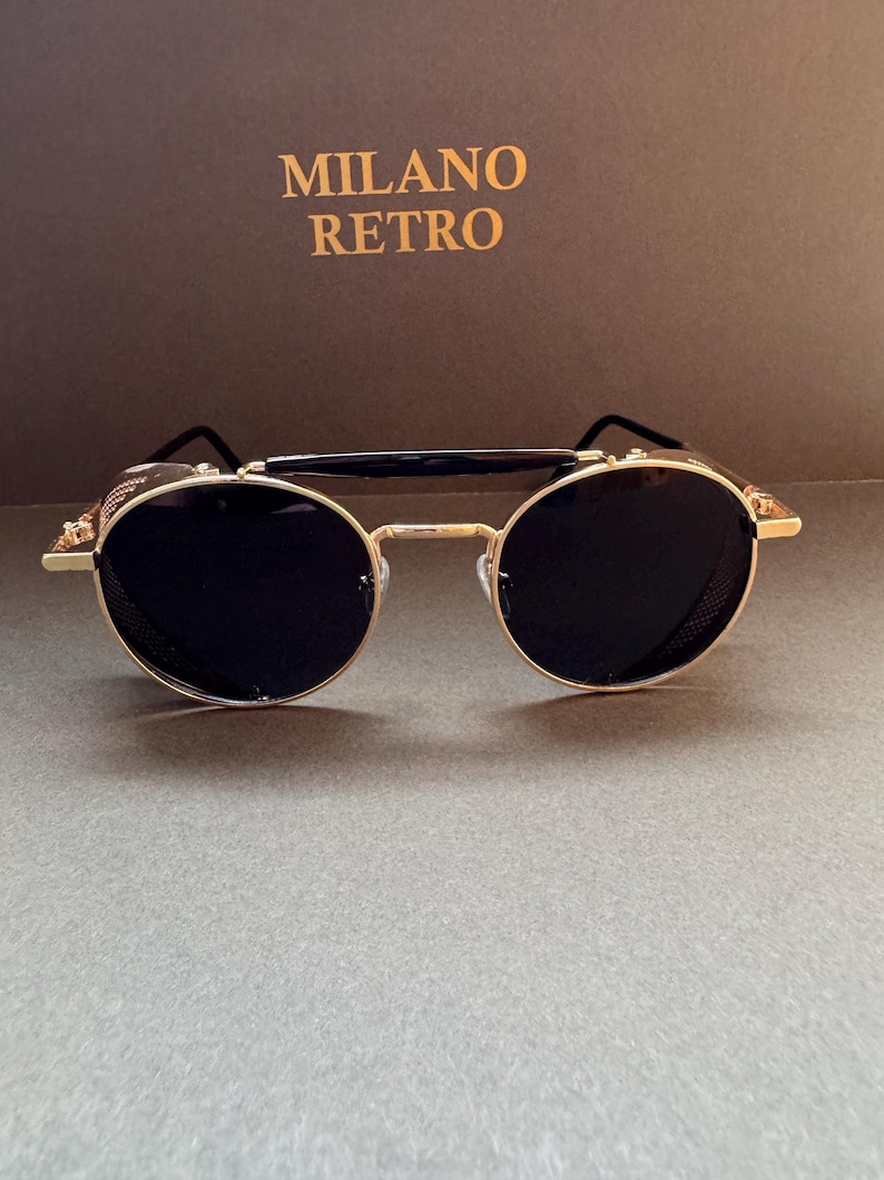 May include: Gold-rimmed sunglasses with round black lenses. The sunglasses have a double bridge and side shields. The text "MILANO RETRO" is in gold lettering on a brown background.