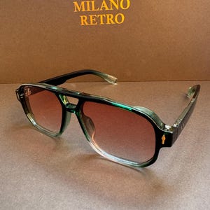 May include: Green and black aviator sunglasses with gradient brown lenses. The sunglasses have a double bridge and a retro design. The text "MILANO RETRO" is in gold letters in the background.