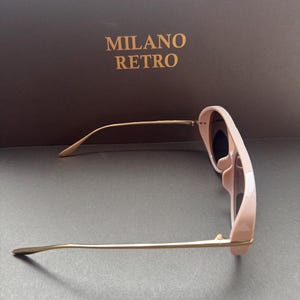 May include: Pale pink sunglasses with gold-tone metal arms. The sunglasses have a rounded frame and dark lenses. The text "MILANO RETRO" is printed in gold on a brown background.