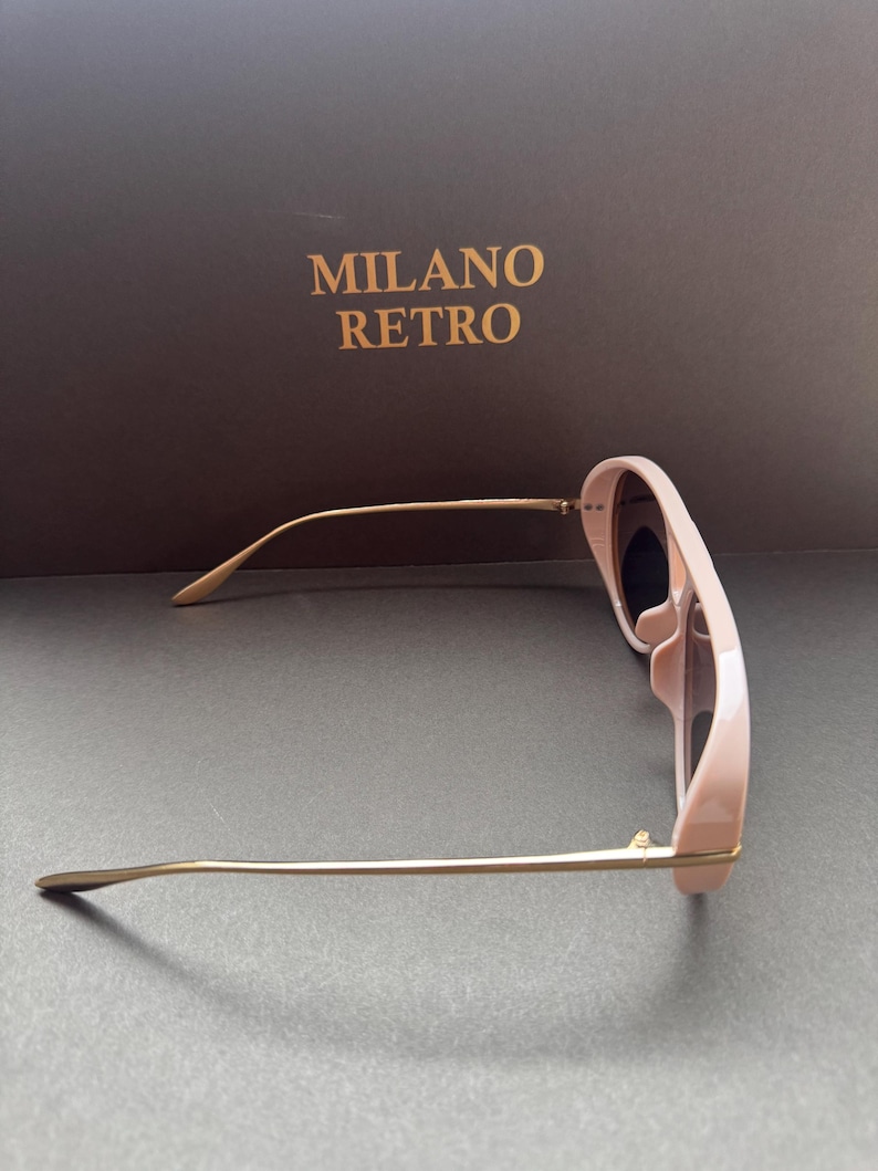 May include: Pale pink sunglasses with gold-tone metal arms. The sunglasses have a rounded frame and dark lenses. The text "MILANO RETRO" is printed in gold on a brown background.