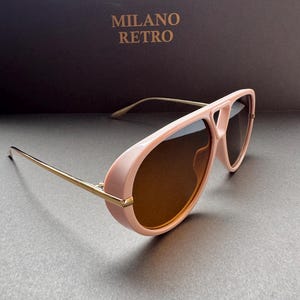 May include: Pink aviator-style sunglasses with gold-colored arms and brown lenses. The sunglasses have a thick frame and a bridge that connects the lenses. The text "MILANO RETRO" is visible in the background.