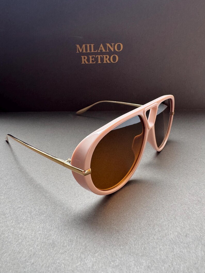 May include: Pink aviator-style sunglasses with gold-colored arms and brown lenses. The sunglasses have a thick frame and a bridge that connects the lenses. The text "MILANO RETRO" is visible in the background.