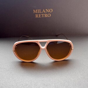 May include: Pink aviator-style sunglasses with brown lenses. The sunglasses have a distinctive bridge design and gold-colored accents. The text "MILANO RETRO" is visible in the background.