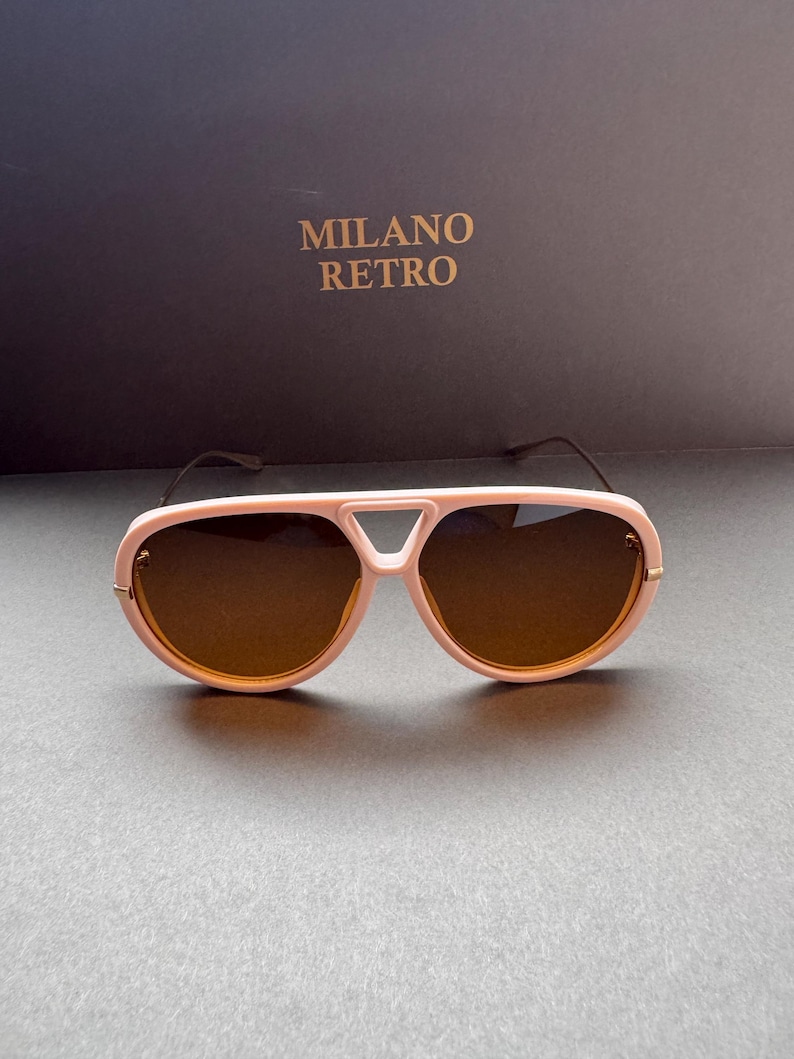 May include: Pink aviator-style sunglasses with brown lenses. The sunglasses have a distinctive bridge design and gold-colored accents. The text "MILANO RETRO" is visible in the background.