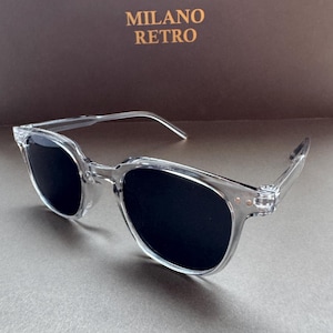 Op de afbeelding: Zonnebril met helder montuur en donkere glazen. De zonnebril heeft een vierkante vorm en staat tegen een bruine achtergrond met de tekst "MILANO RETRO" in gouden letters.