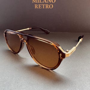 May include: Brown aviator sunglasses with gold-coloured accents. The sunglasses have a double bridge and a retro design. The text "MILANO RETRO" is visible in gold lettering on the background.