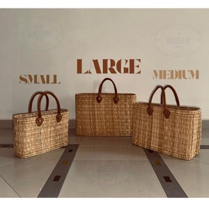 May include: Three woven straw tote bags in different sizes: small, medium, and large. Each bag has brown leather handles and trim. The bags are set against a neutral background with size labels above them.