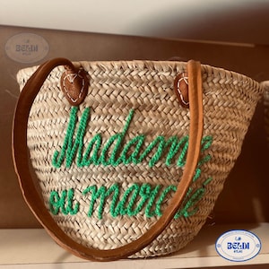 May include: Woven straw tote bag with brown leather handles and green embroidered text that reads "Madame ou morte". The bag has a rounded shape and is suitable for carrying items.