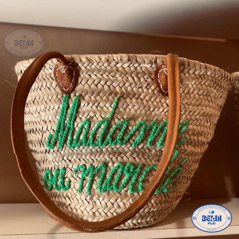 May include: Woven straw tote bag with brown leather handles and green embroidered text that reads "Madame ou morte". The bag has a rounded shape and is suitable for carrying items.
