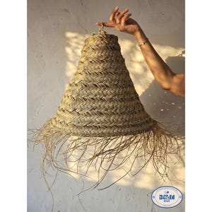 May include: A woven, conical lampshade made of natural fibers. The lampshade has a wide, fringed bottom and is held up by a hand. The shade is a light tan color and appears to be made of straw or similar material.