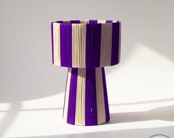 Puede incluir: Una lámpara decorativa con una pantalla cilíndrica y una base de pedestal. La lámpara presenta rayas verticales de hilo morado y crema. La lámpara es un artículo de decoración del hogar único.