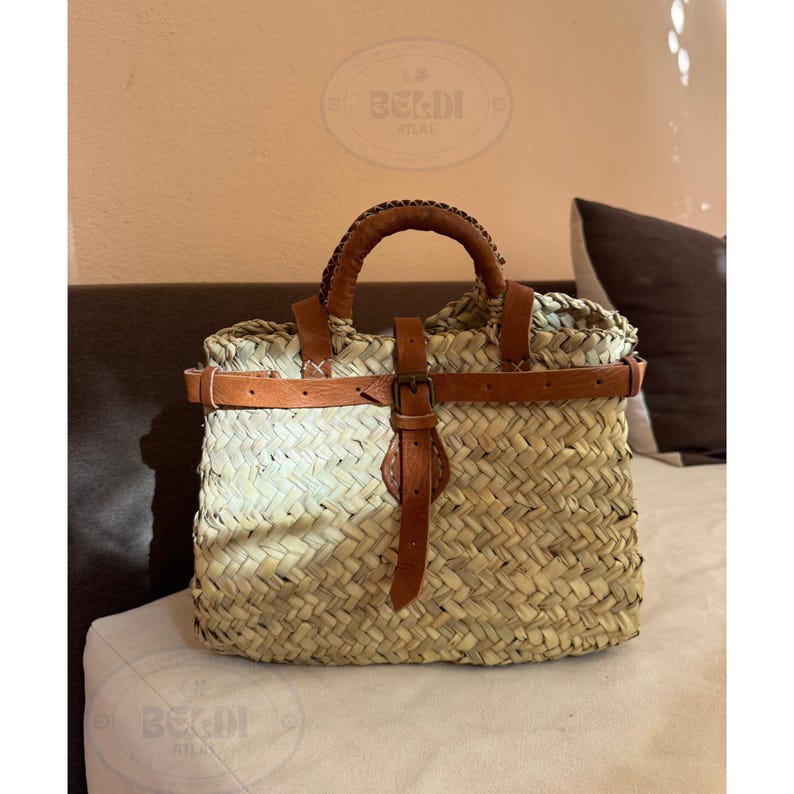 May include: Woven straw tote bag with brown leather accents. The bag features a rectangular shape with a woven pattern, a brown leather handle, and a leather strap with a buckle closure. The bag has the text "BELDI ATLAS" on it.